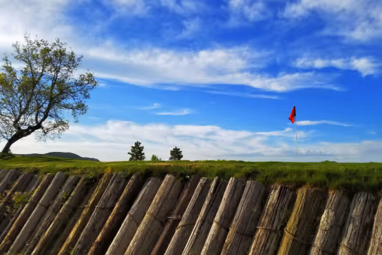 Red golf flag on a green hill with a dramatic wooden retaining wall and a bright blue sky.