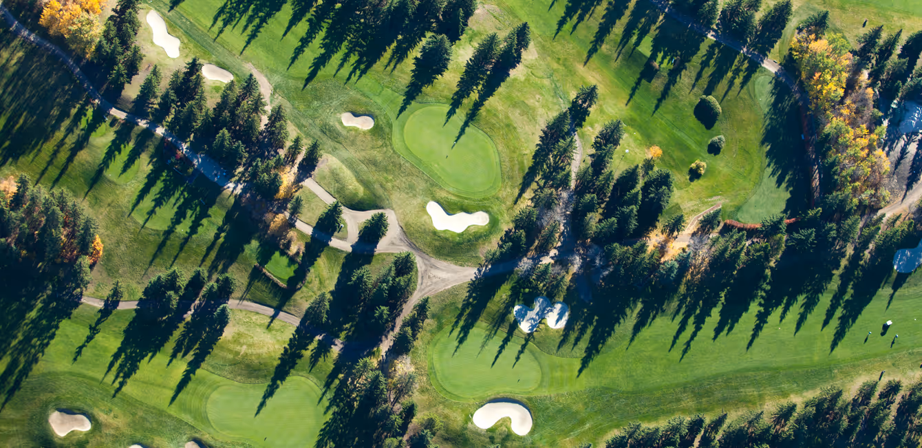 Aerial view of a tree-lined golf course with putting greens, sand bunkers, and winding paths casting long shadows.