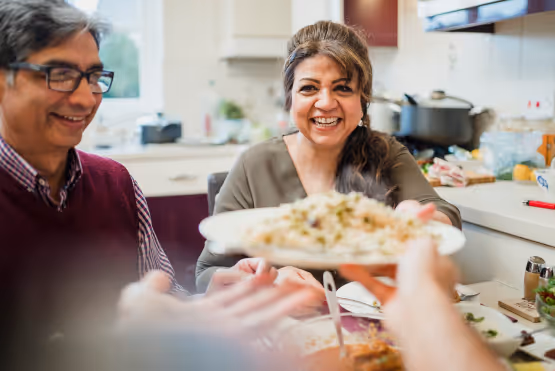 A smiling woman passing a plate of food across the table during a family meal in a cozy kitchen.