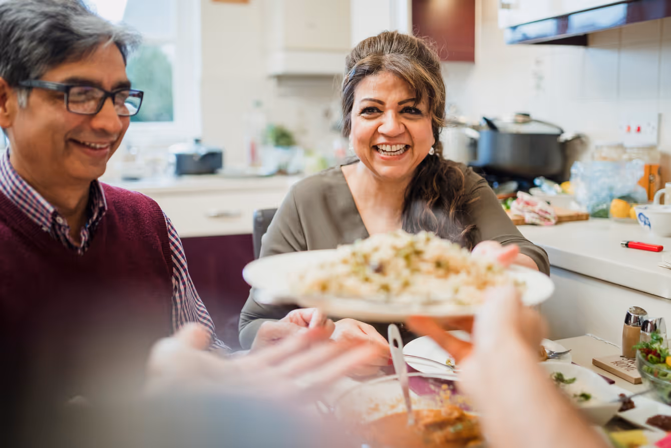 A smiling woman passing a plate of food during a family meal in a cozy kitchen, symbolizing warmth and hospitality.