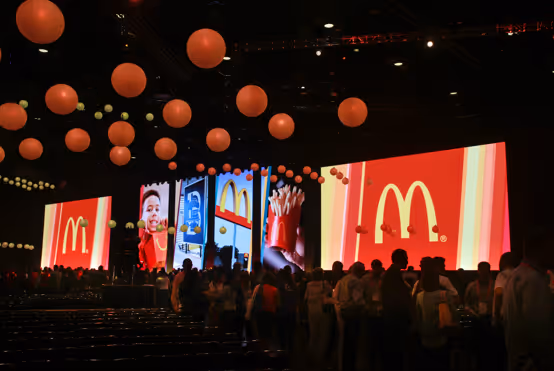 Large McDonald’s convention stage with multiple screens displaying the golden arches logo and food images, with attendees gathered in front under hanging lights.