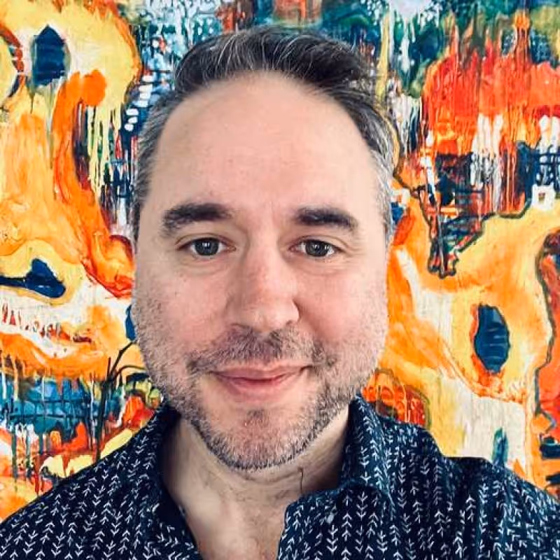 Close-up of a man with short dark hair and light stubble smiling in front of a colorful abstract painting.