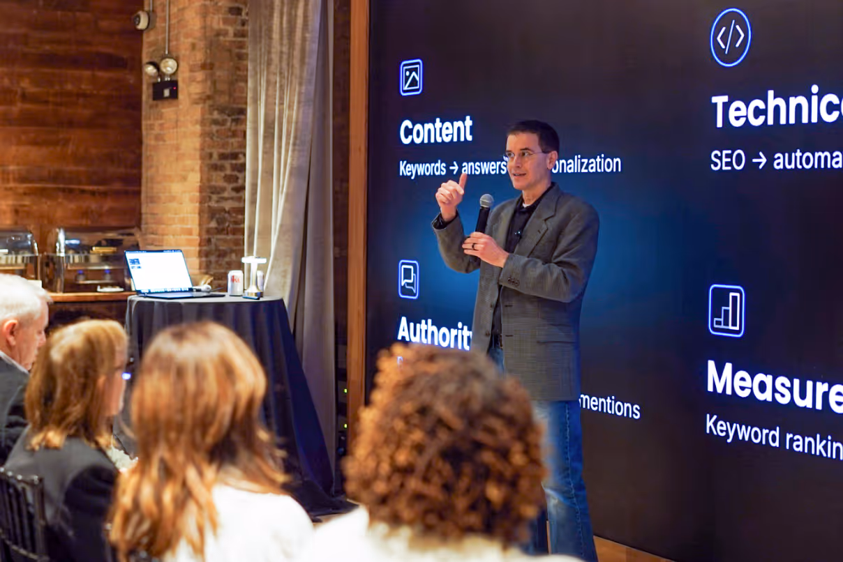 Man in gray blazer giving a presentation with a microphone in front of a large screen showing content strategy terms like Content, Authority, Technical, and Measurement.
