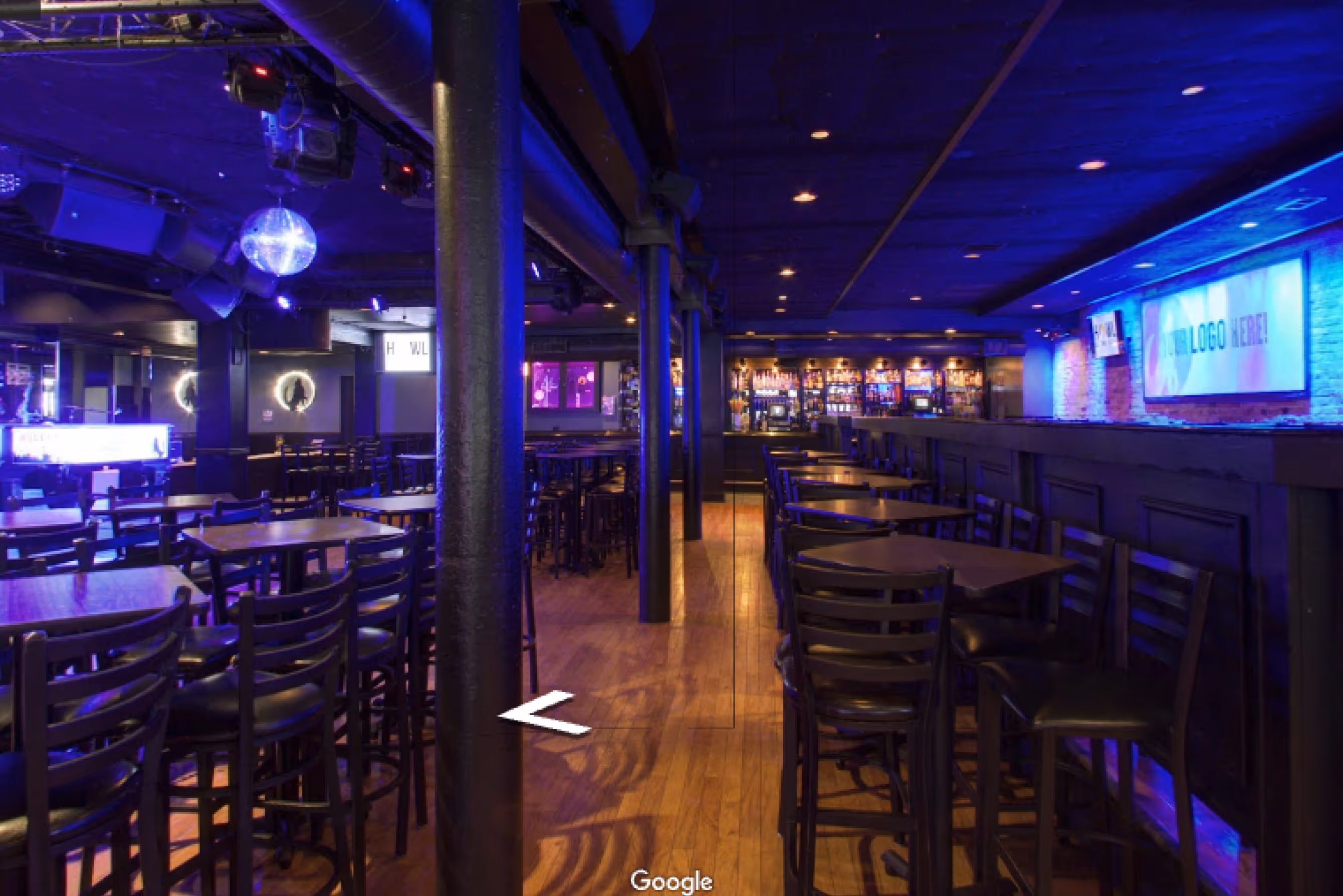 Empty bar and restaurant interior with rows of black chairs and tables, a wooden floor, and colorful purple and blue lighting.