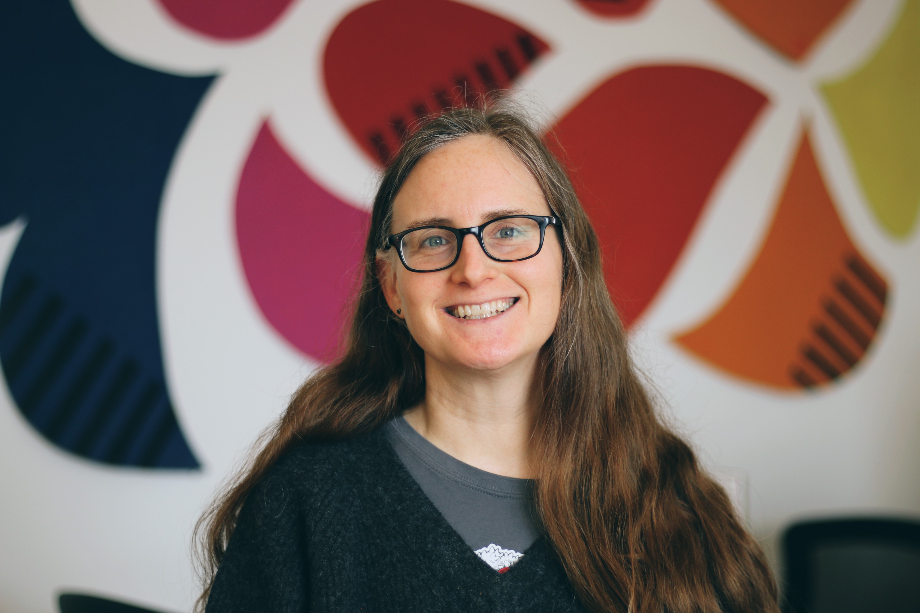 Photo of a person with glasses smiling at the camera against a wall with a colorful mural.