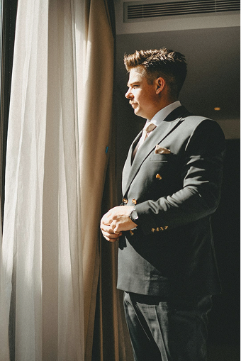 Man in a dark suit looking thoughtfully out a window with sunlight streaming in.
