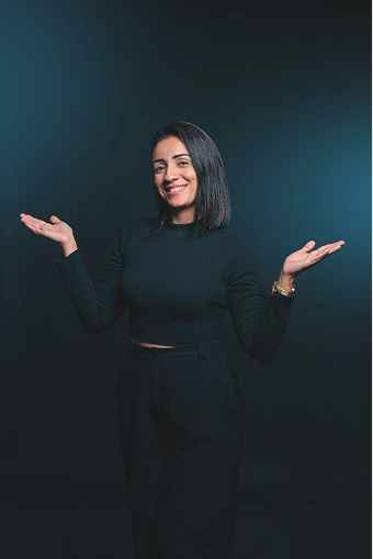Smiling woman with shoulder-length black hair wearing a black long-sleeve crop top and high-waist pants, posing with arms raised and palms up against a dark background.