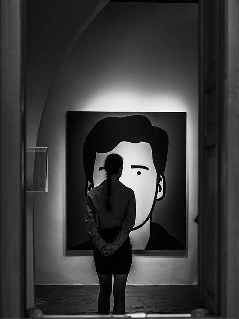 Person with braided hair standing with hands behind back, viewing a large backlit minimalist portrait of a face in a gallery.