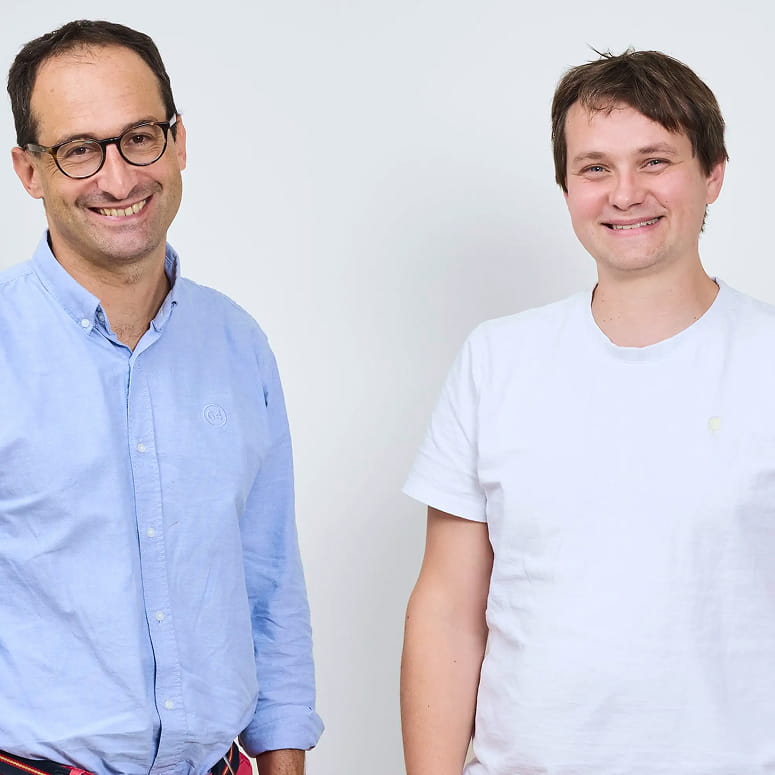 Two men smiling against a plain white background, one wearing glasses and a light blue button-up shirt, the other in a white t-shirt.