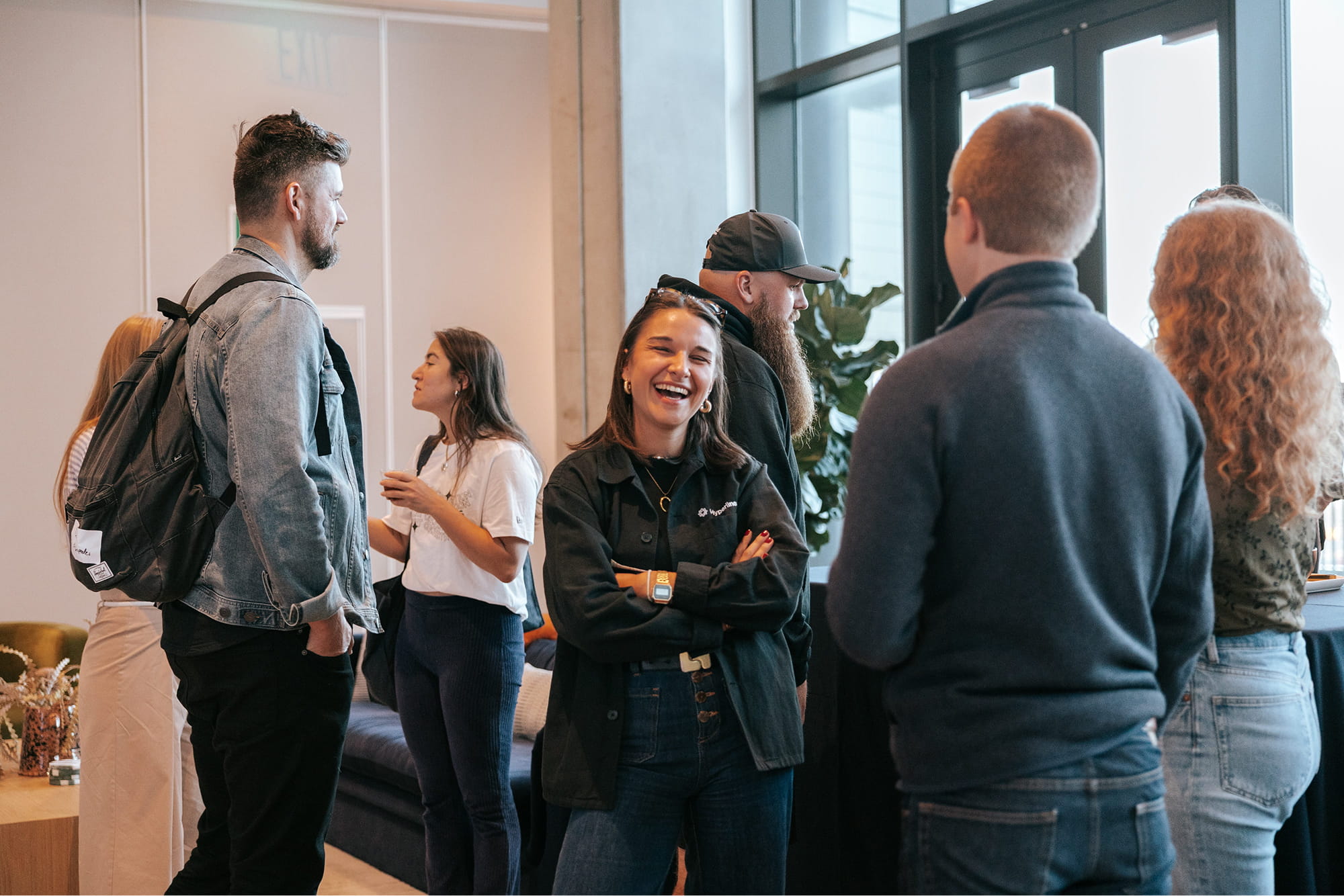 Group of young adults socializing indoors, with one woman in the center laughing and others engaged in conversation.