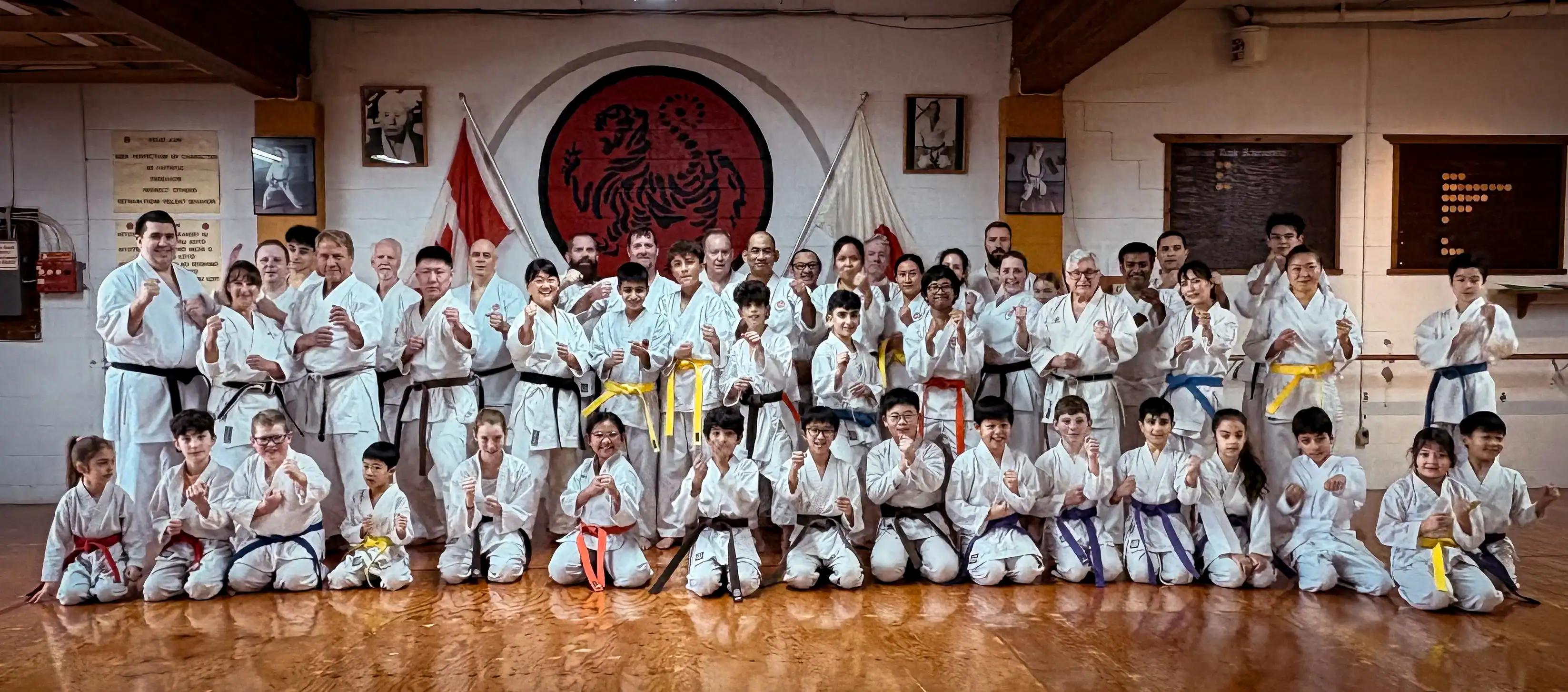 2025 group photo of Port Moody Shotokan Karate Club in Port Moody, BC, with students in white gi and colored belts posing in the dojo under the Shotokan tiger emblem, Dojo Kun, and flags.