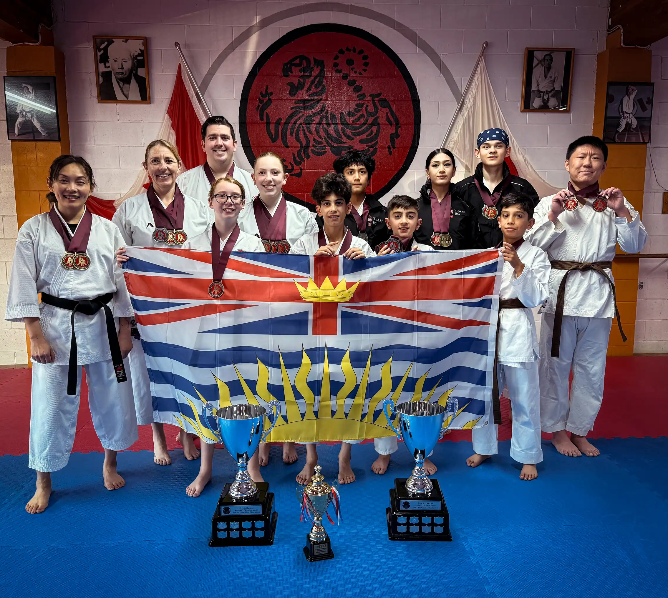 Port Moody Shotokan Karate Club young competitors posing with medals, trophies, and British Columbia flag in dojo featuring Shotokan tiger emblem, December 2025.