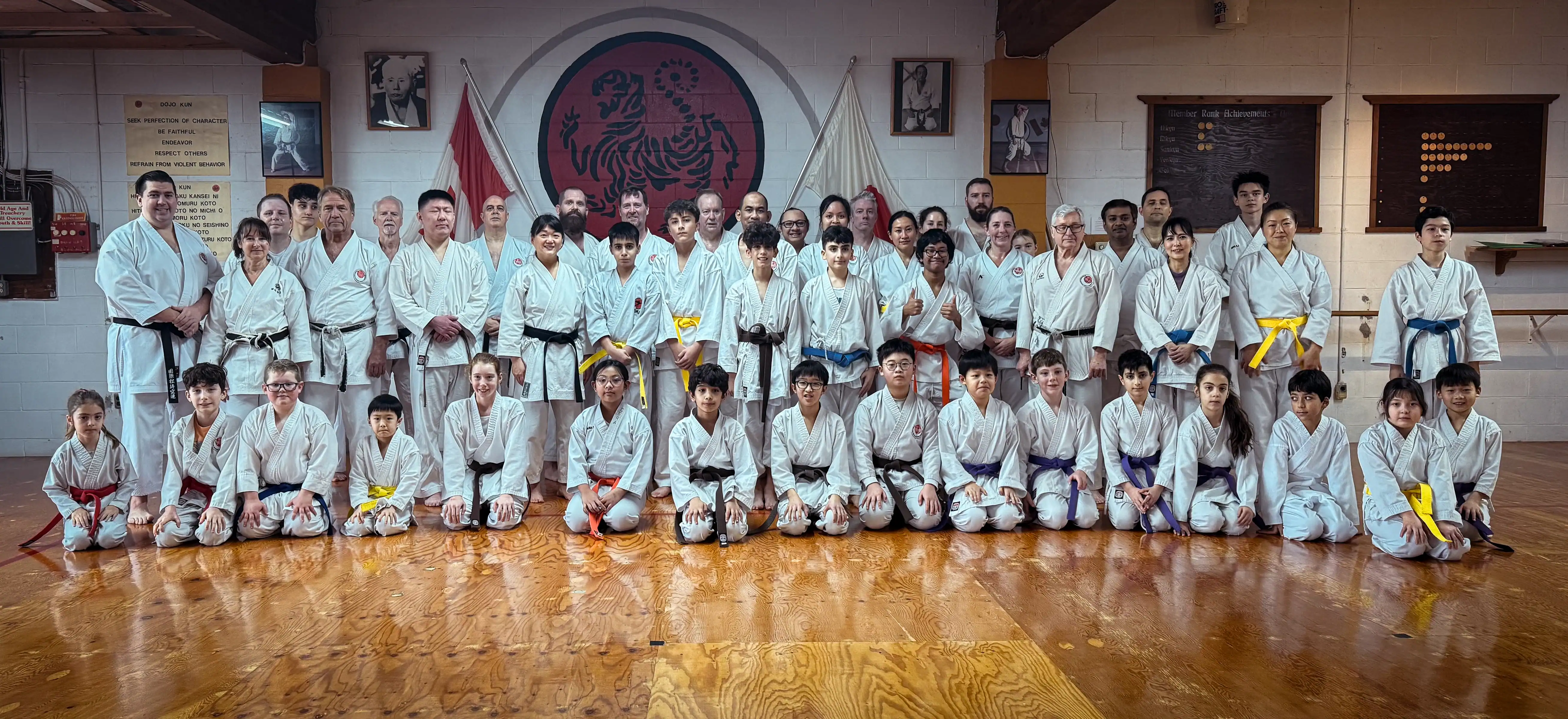 Group photo of Port Moody Shotokan Karate Club members in Port Moody, British Columbia, posing in traditional white karate uniforms with colored belts in a dojo featuring the Shotokan tiger emblem, Dojo Kun, and Japanese flags.