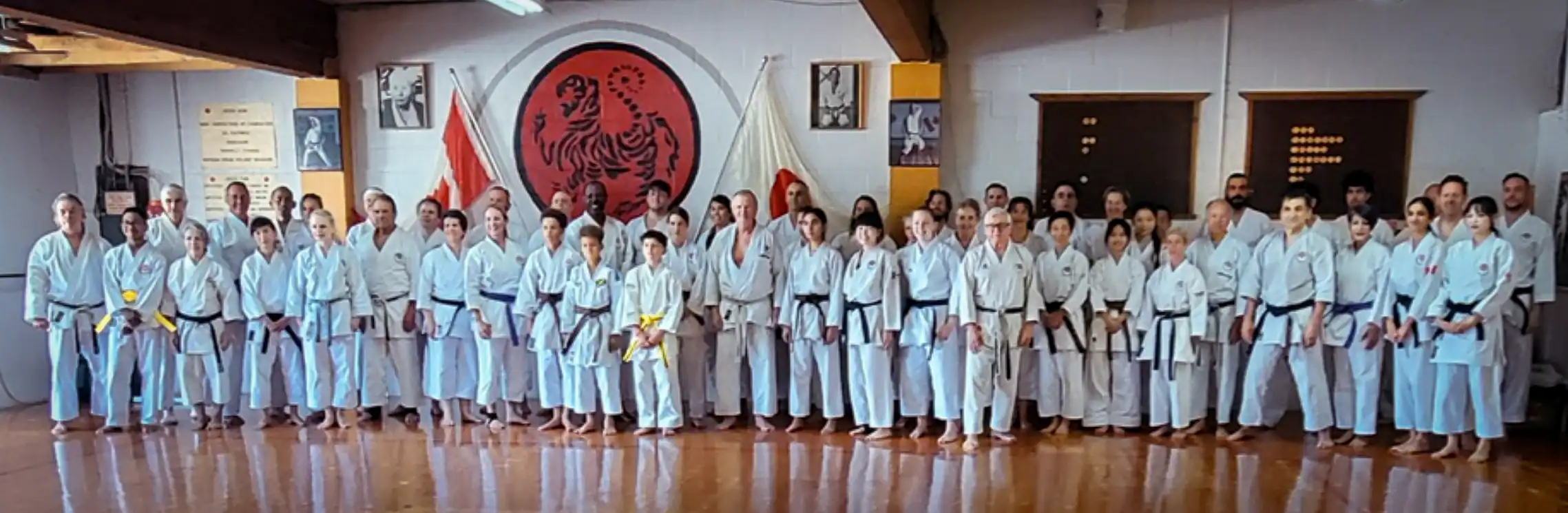 Large group photo of Port Moody Shotokan Karate members of all ages and ranks in white gi, standing in the dojo under the Shotokan tiger emblem and Dojo Kun, 2025.