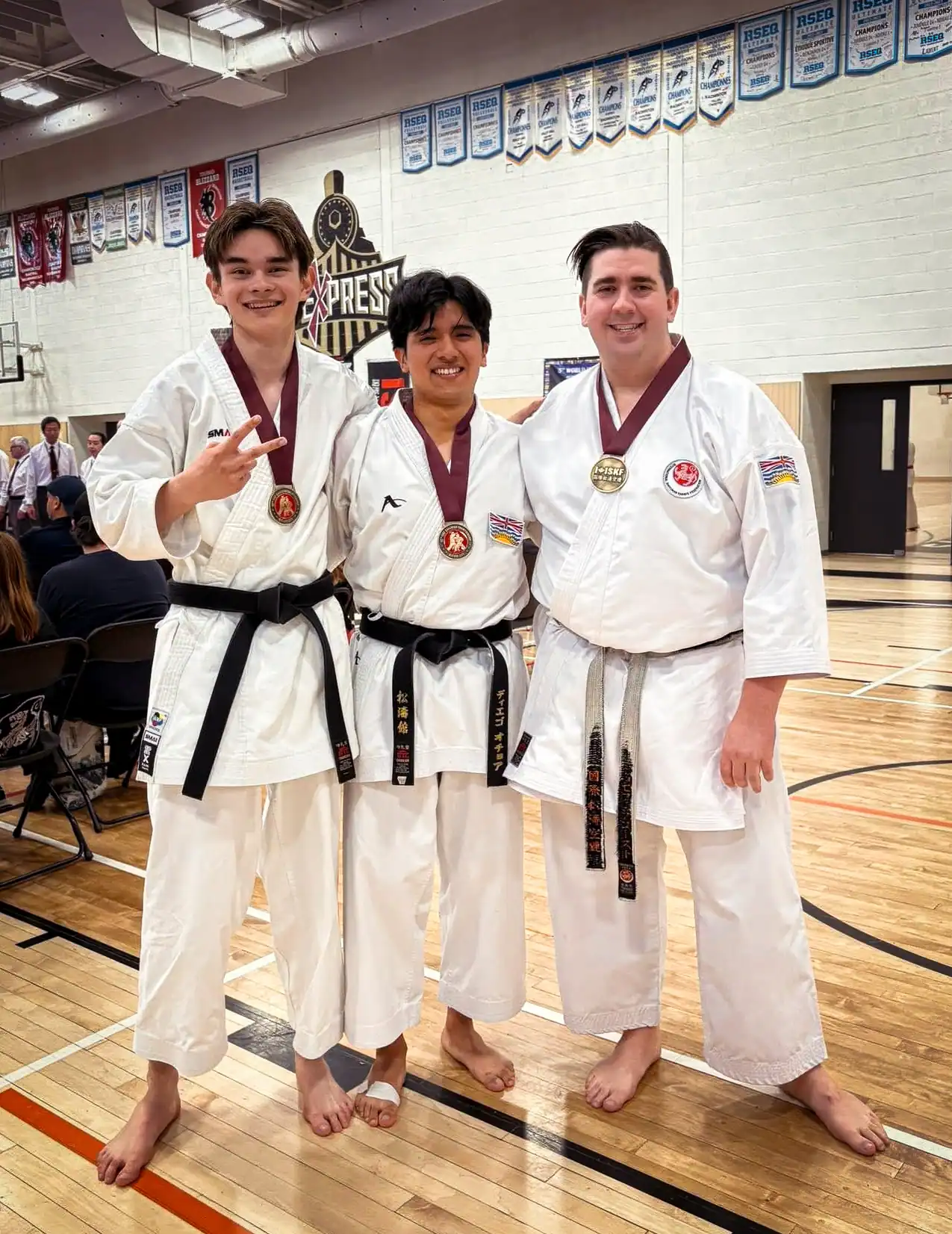 Port Moody Shotokan Karate Club black belts Charlie, Diego, and Joseph wearing medals after a 2025 competition, posing in gymnasium with championship banners.