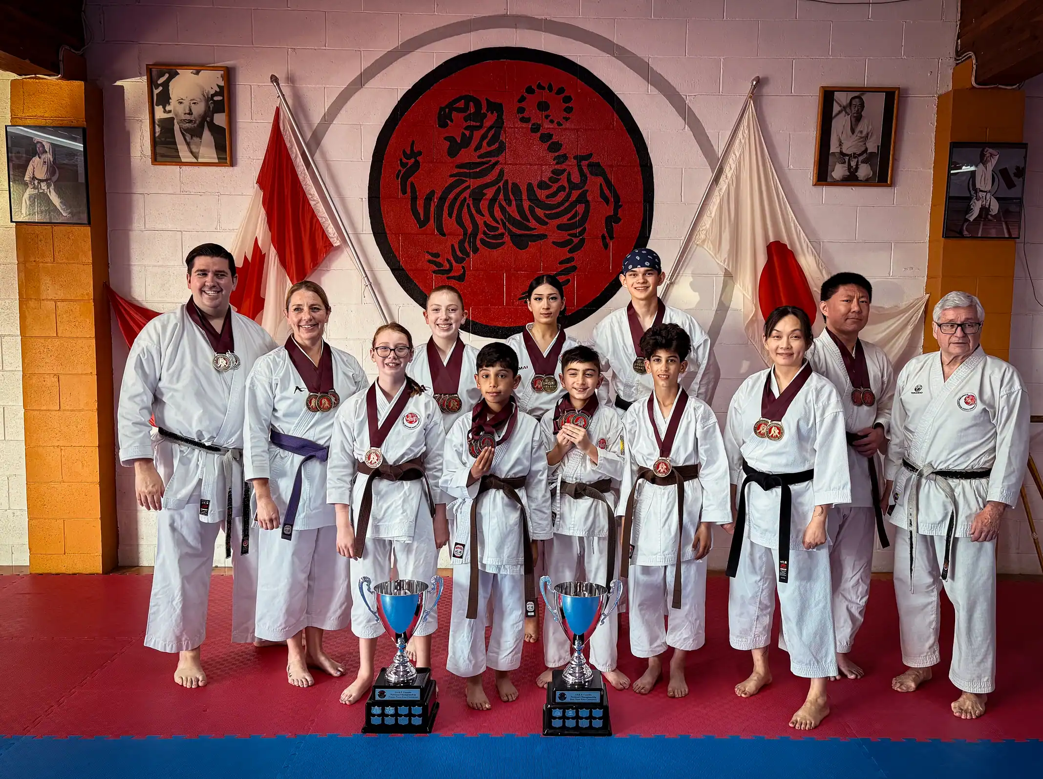 Port Moody Shotokan Karate 2025 tournament medal winners group photo: young and senior students in white gi with medals and trophies, standing in the traditional dojo under the Shotokan tiger logo and flags of Canada and Japan.