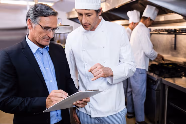 Chef in white uniform discussing with a man in a suit holding a clipboard in a restaurant kitchen.