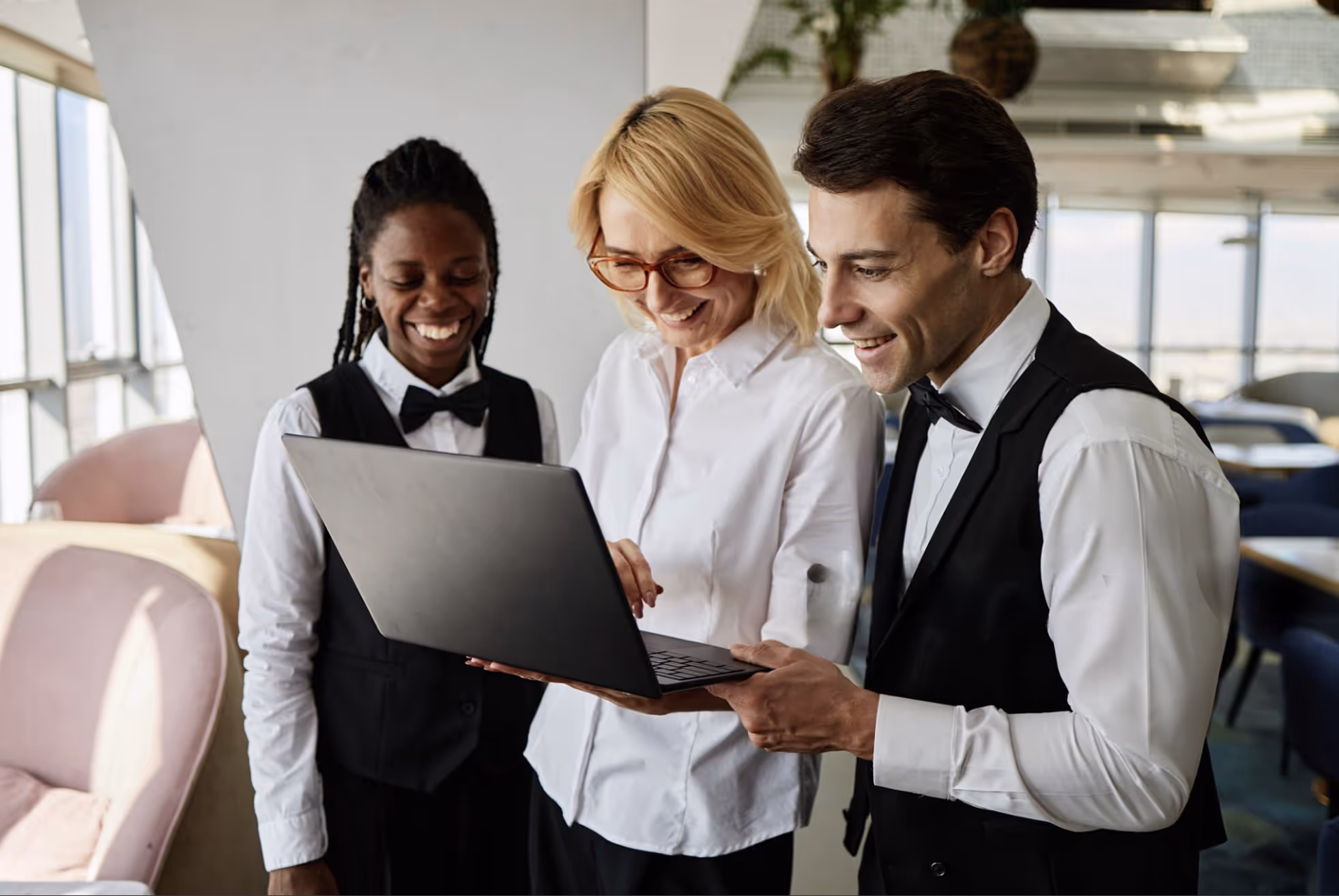 Three smiling restaurant staff in white shirts and black vests looking at a laptop together indoors.