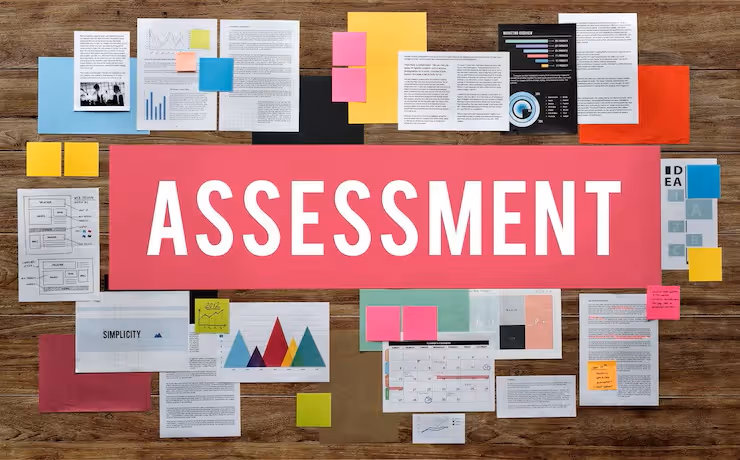 Wooden table with various documents, charts, and sticky notes arranged around a large red paper displaying the word 'ASSESSMENT' in white letters.