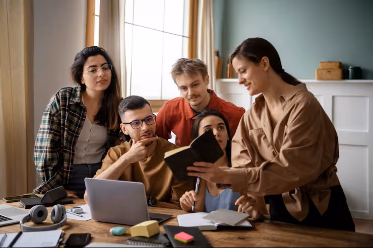 A group of five young adults gathered around a table studying together, with one woman holding and showing a book to the others.