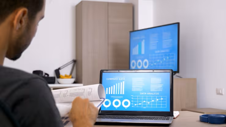 Person reviewing printed charts while a laptop and a large monitor display company data graphs and data analysis charts.