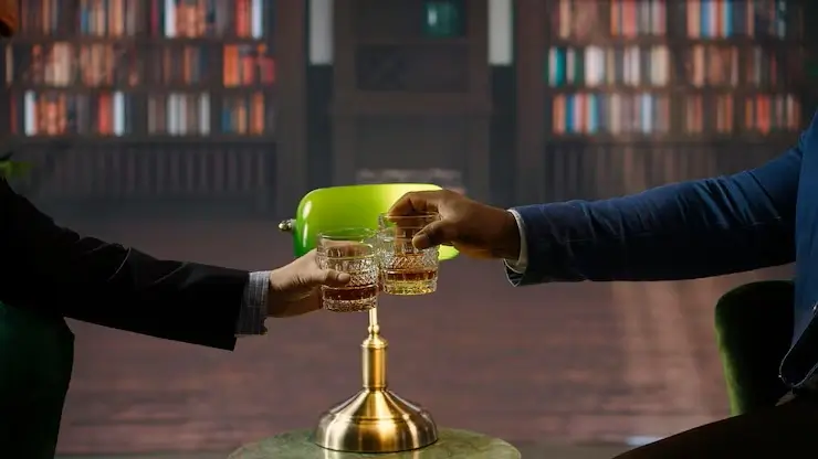 Two people clinking glasses of whiskey in a dimly lit library with a green banker’s lamp on a table between them.