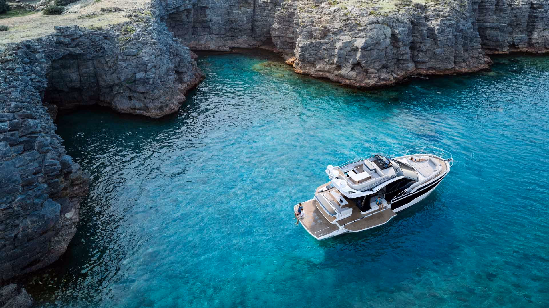 Galeon 420 FLY motor yacht anchored in a turquoise cove beside limestone cliffs, with guests relaxing on the swim platform and foredeck