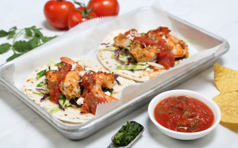 Shrimp tacos topped with chunky tomato salsa, served alongside fresh herb garnish and tortilla chips.