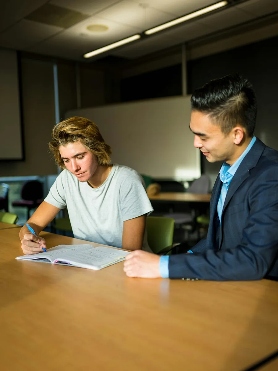 A tutor smiling as a student writes notes in a notebook at a classroom table.