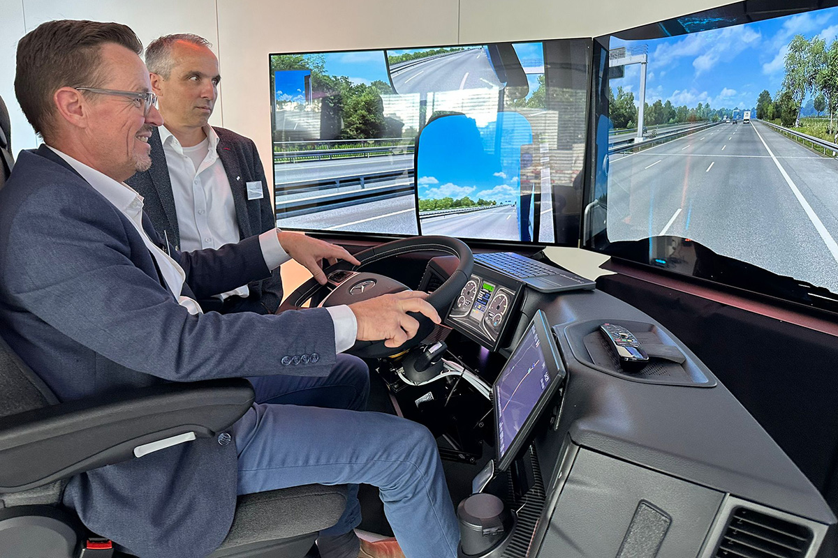 Mann testet Lkw-Fahrsimulator mit großem Bildschirm und Lenkrad, daneben beobachtet ein weiterer Mann.