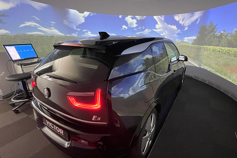 Schwarzer BMW i3 steht in einem Fahrsimulator mit virtueller Landschaft auf Bildschirmwänden.