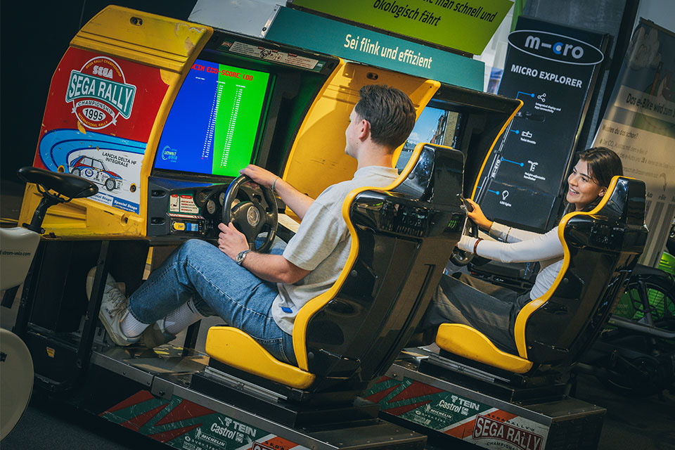 Zwei Personen spielen SEGA Rally Arcade-Rennspiel auf einem Simulator mit Lenkrad und Pedalen.