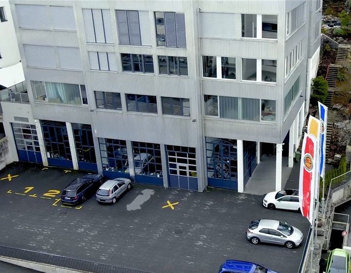 Mehrstöckiges Gebäude mit blauen Türen, Autos auf Parkplatz und drei Flaggen neben Eingang.