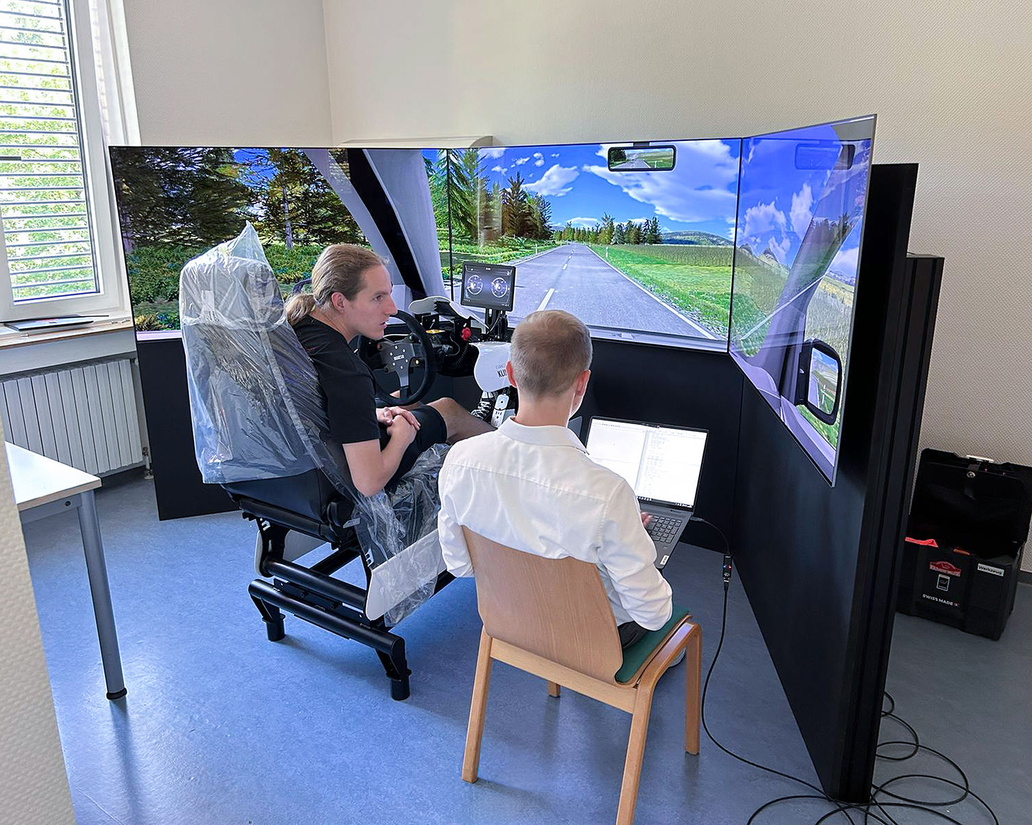Deux hommes testent un simulateur de conduite avec trois écrans et un volant dans un bureau.