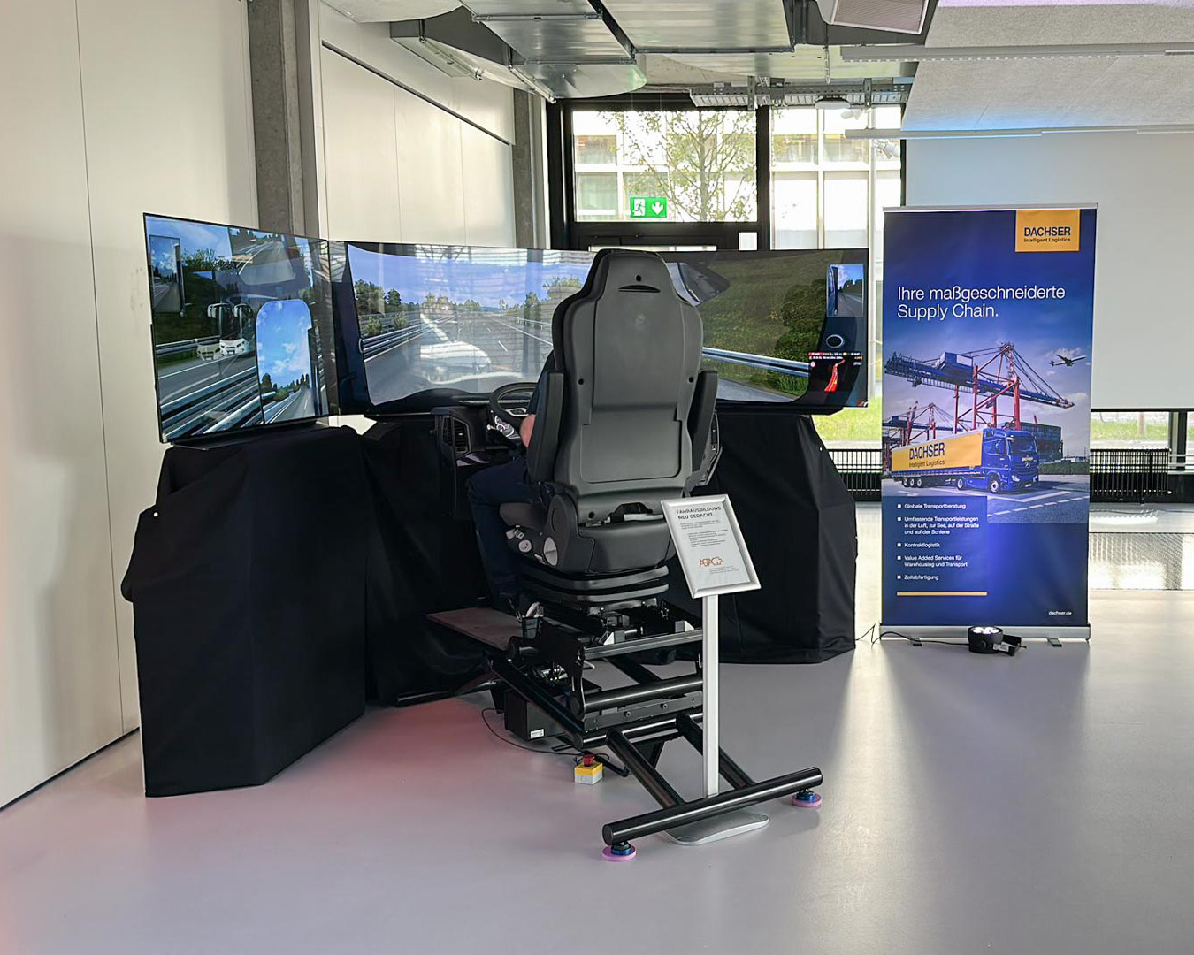 LKW-Fahrsimulator mit mehreren Bildschirmen und Sitz, im Raum neben einem Werbebanner von Dachser.