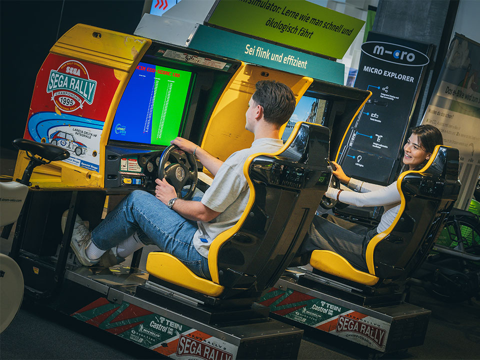 Zwei Personen spielen auf gelben SEGA Rally Fahrsimulatoren nebeneinander in einer Ausstellung.