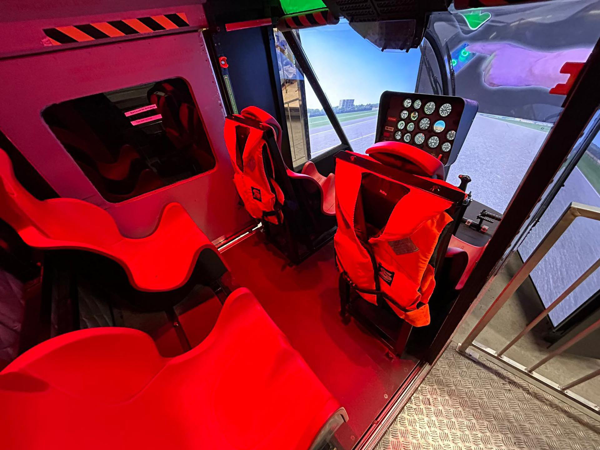 Helikoptercockpit mit roten Sitzen, Steuerpult und Außensicht auf Startbahn und Himmel.
