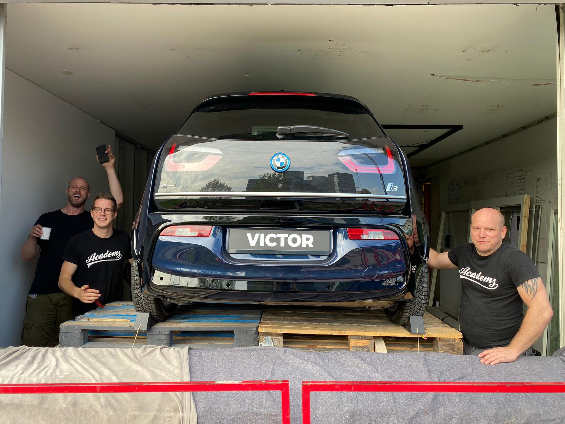 Vue arrière d'une BMW i3 noire sur des palettes en bois, avec trois hommes souriants à côté d'eux.