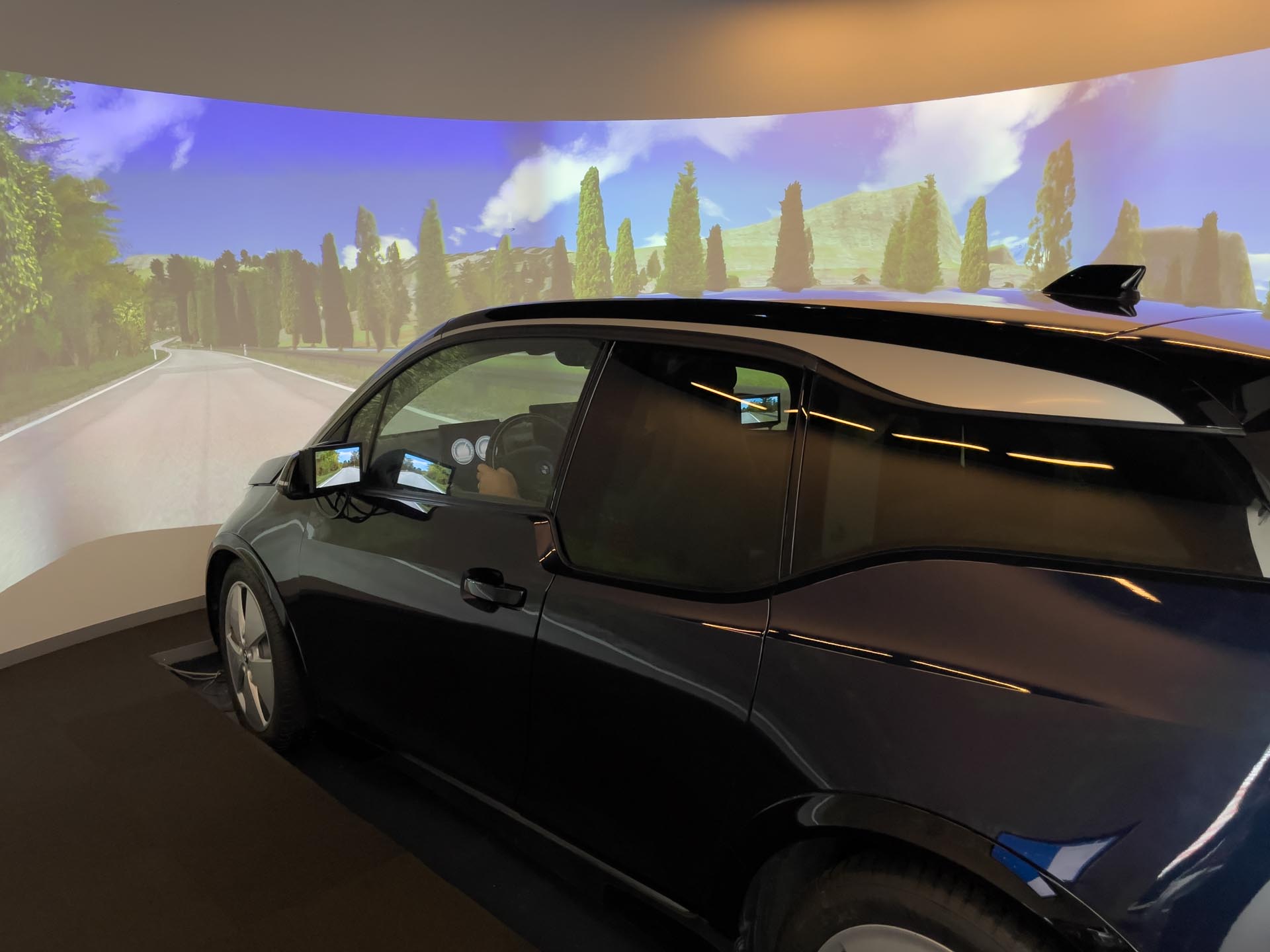 Conducteur en voiture noire conduisant dans une simulation de conduite avec projection de paysage tout autour.
