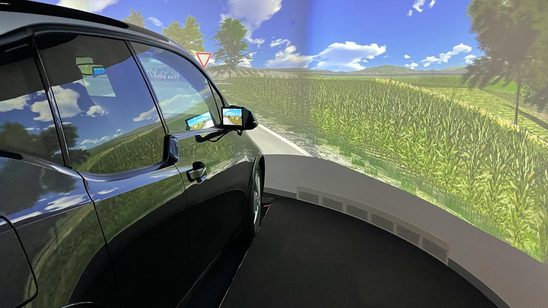 Une voiture dans une simulation de conduite montre une route rurale avec des champs et un ciel bleu.