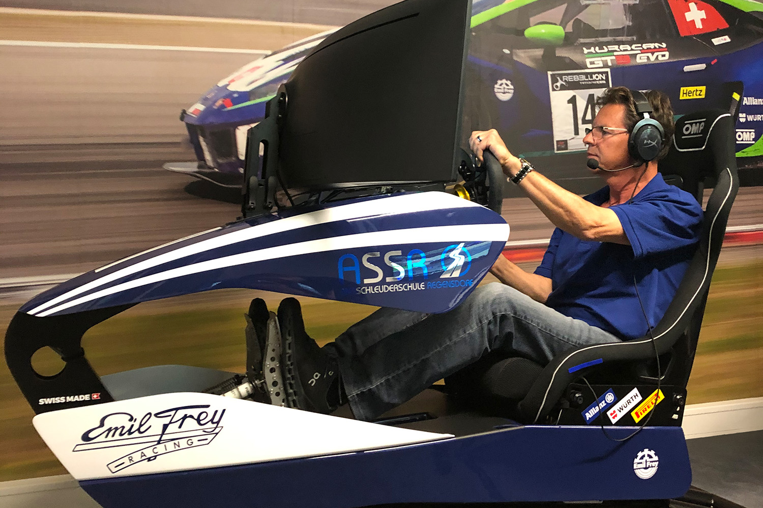 Man sitting in a closed racing simulator with steering wheel and headset in front of racing car mural.