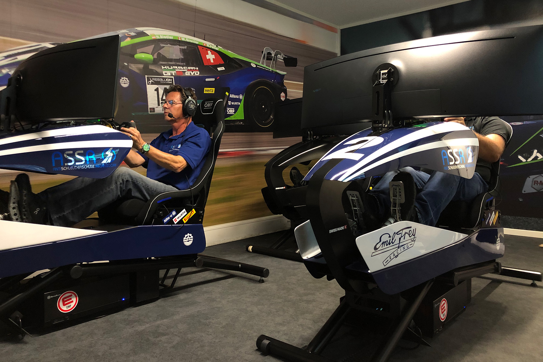 Two people drive in racing simulators with large screens and headphones in the interior.