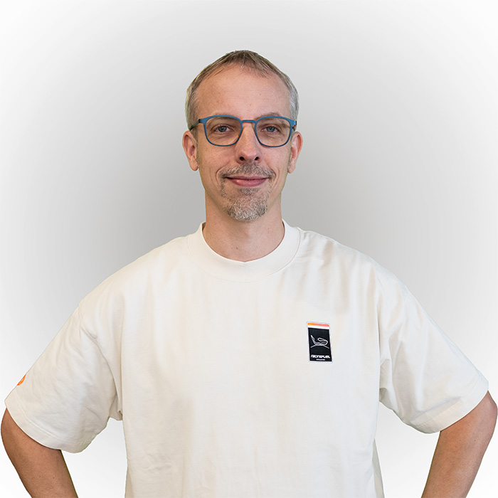 Man wearing blue glasses wears a white T-shirt and smiles slightly against a light gray background.
