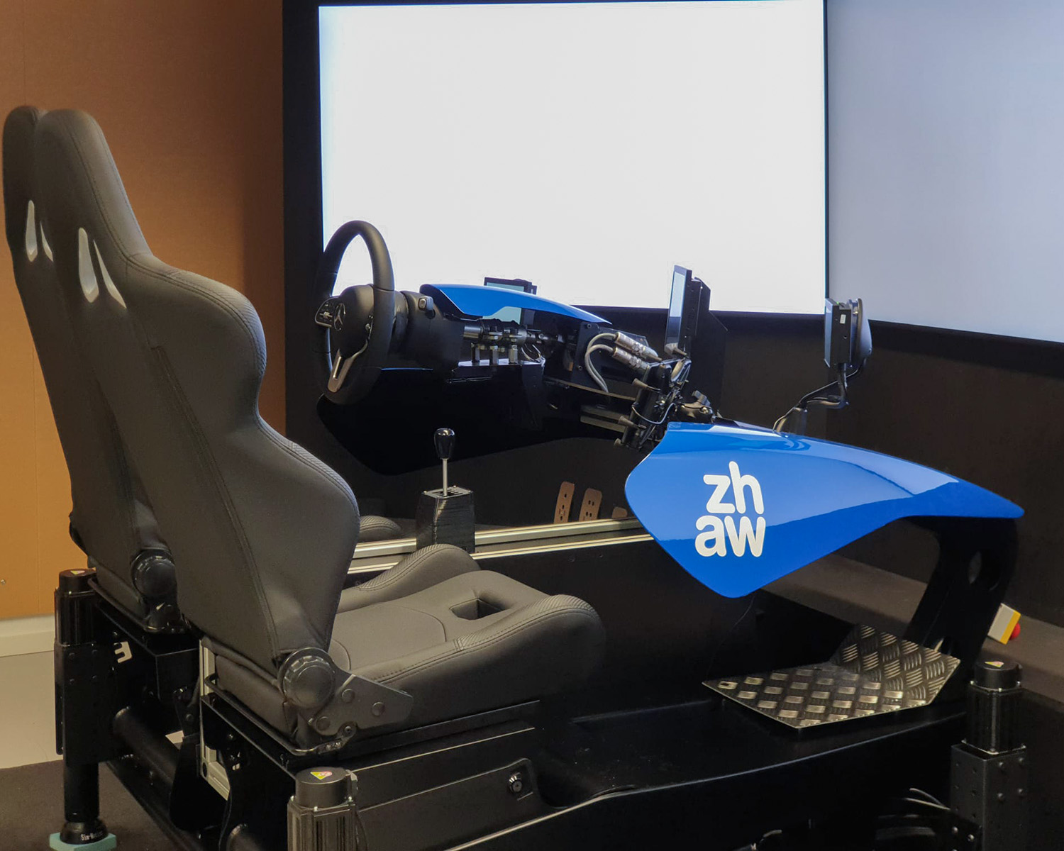 Simulateur de conduite avec deux sièges noirs et garniture bleue avec le logo ZHAW devant un grand écran.