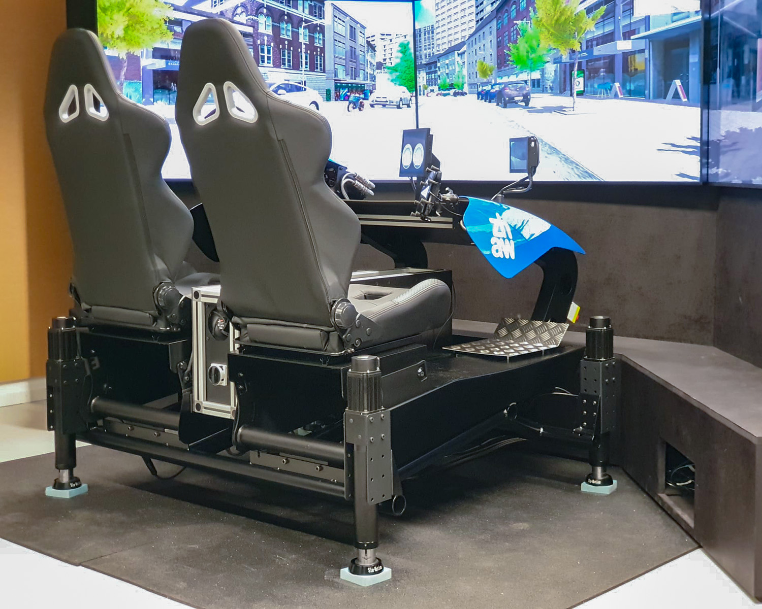 Two seats with steering wheel and screens in front of a large simulation of a city scene.
