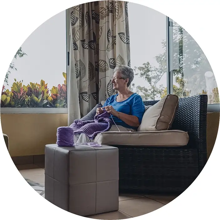 Ältere Frau sitzt auf einem Sofa, strickt einen Pullover und schaut zufrieden aus dem Fenster – häusliche Pflege und selbstbestimmtes Leben im Alter
