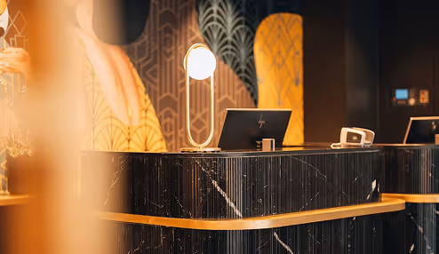 Modern hotel reception desk with a black marble-like front, lit decorative lamp, computer, and elegant patterned wall in the background.