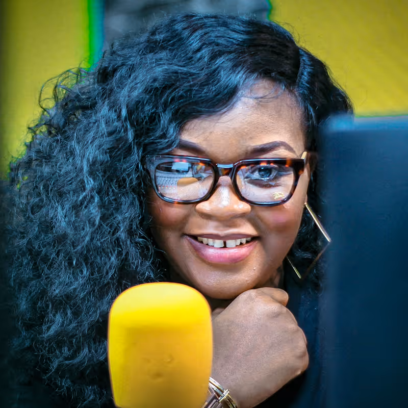 Smiling woman with glasses speaking into a yellow microphone in a recording studio.