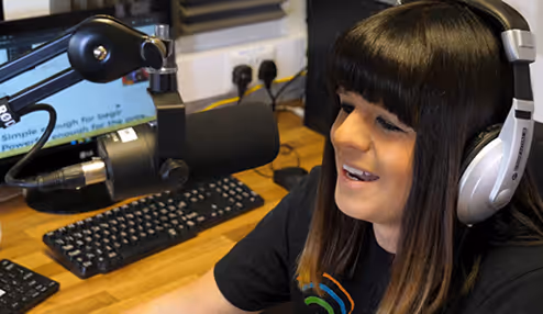Radio Alty. Person with dark hair wearing headphones speaking into a microphone in a radio studio.
