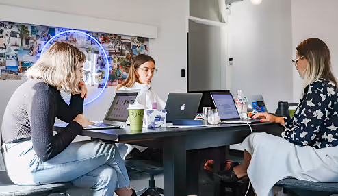 Three women working on laptops around a black table in a modern office setting with a neon sign on the wall.